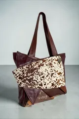 Cartera de cuero marrón estilo tote bag, con diseño en patchwork y bolsillo frontal de pelo con estampado animal print tipo vaca. Cuenta con doble asa de hombro y detalles metálicos.