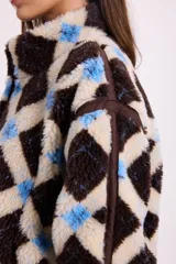 Campera de corderito sintético con diseño a cuadros en tonos marrones y negros. Presenta cuello alto, cierre frontal completo y detalles de paneles en color marrón liso. El diseño es de silueta holgada, largo tipo crop y cuenta con un cordón elástico ajustable en el ruedo.