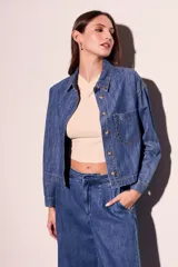 Camisa de jean azul de corte recto, con cuello clásico, cierre frontal con botones marrones, bolsillo de parche en el pecho y mangas largas con puños abotonados.