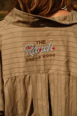 Camisa color arena a rayas verticales finas, de corte amplio, con cuello camisero, cierre frontal con botones, bolsillo de parche en el pecho con bordado de caballo rojo, mangas largas con puños elásticos y cinturón ancho de tela con flecos.