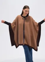 Poncho de tejido grueso color camel, con bordes y mangas en color marrón oscuro. Cierre frontal con broche rectangular negro.