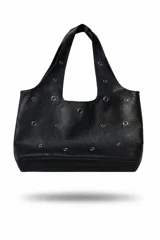 Cartera tipo tote de eco cuero negro con textura granulada, decorada con múltiples ojalillos metálicos plateados distribuidos en el frente y las asas. Cuenta con cierre de botón y forro interior con bolsillo.