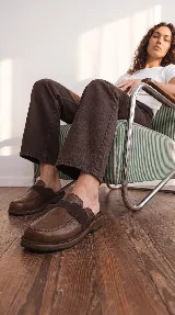 Zueco de cuero marrón, con diseño inspirado en mocasines, tira elástica en el empeine y suela plana confortable.