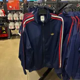 Campera deportiva Adidas Originals de color azul marino con cierre frontal. Presenta el logo del trébol bordado en dorado en el pecho y las icónicas tres tiras en rojo y blanco a lo largo de las mangas.
