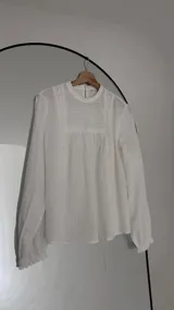 Blusa blanca de corte holgado con cuello redondo, mangas largas con puños elásticos y detalles de puntillas verticales en el frente.