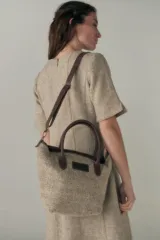 Cartera tipo bolso de mano confeccionada en lana fina sin teñir, con detalles en cuero marrón oscuro en las asas y correa ajustable. Incluye cierre superior y etiqueta de cuero con logo.