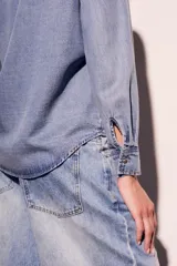 Camisa de denim celeste con cuello clásico, cierre frontal con botones a presión, dos bolsillos con solapa abotonada en el pecho y mangas largas con puños abotonados.