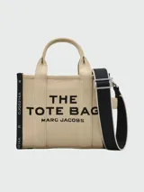 Cartera tipo tote bag de lona color beige, con diseño estructurado y asas superiores. Presenta el logo de la marca estampado en la parte frontal y detalles de cintas laterales con inscripciones.