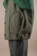 Chaleco largo acolchado color verde oliva, reversible con un lado de piel sintética y otro de pelo sintético. Tiene cuello en V, cierre con botones a presión y bolsillos laterales. Silueta amplia y relajada.