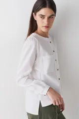 Blusa de lino color blanco, con cuello redondo, cierre frontal con botones de madera y dos bolsillos delanteros. Presenta mangas largas con puños abotonados y costuras a la vista en contraste.