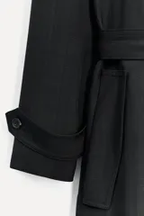 Trench negro largo con cuello solapa, manga larga con trabilla y botón, bolsillos delanteros, cinturón ajustable con lazadas, abertura en la espalda y cierre frontal con botones ocultos por solapa.