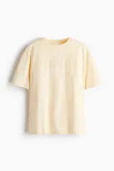 Remera oversized de punto de algodón color amarillo claro, con cuello redondo, mangas cortas y hombros caídos. Cuenta con la palabra "ATELIER" bordada en relieve en el frente.