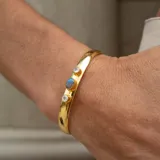 Brazalete rígido dorado con tres piedras incrustadas: una labradorita ovalada en el centro y dos piedras de la luna redondas a los lados.