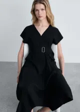 Vestido midi de corte evasé en color negro, con cuello en pico y diseño sin mangas. Presenta un cinturón ajustable con hebilla metálica en la cintura, dos bolsillos con solapa en la parte frontal y un bajo asimétrico.