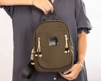 Mochila de nylon color verde oliva, con compartimento principal y bolsillo frontal con cierre metálico. Presenta detalles de cordones decorativos en los tiradores de los cierres, una grifa de goma con el logo de la marca y correas ajustables.