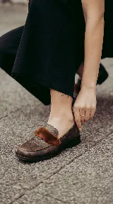Mocasín de cuero con textura gastada, color marrón, con detalle de pelo en la parte superior y suela plana.