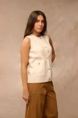 Chaleco de tejido de punto color crema, sin mangas, con cuello redondo y cierre frontal de botones dorados. Cuenta con dos bolsillos frontales aplicados con detalles de botones dorados.
