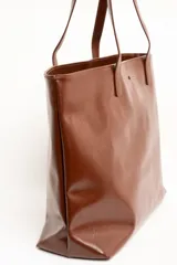 Bolso tipo tote bag color marrón, confeccionado en cuero sintético, con doble asa superior y detalle de placa metálica dorada con logo en la parte frontal.