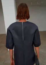 Vestido corto de satén color gris oscuro, con cuello redondo, mangas cortas y diseño recto. Cuenta con cierre de cremallera en la parte posterior y forro interior.