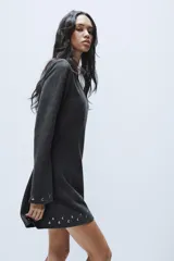 Vestido corto de punto color gris oscuro, con cuello barco y mangas largas acampanadas. Presenta detalles de tachas metálicas aplicadas en el ruedo y en los puños, con un diseño de corte evasé.
