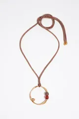 Collar largo con cordón de tela marrón, dije circular metálico dorado con diseño irregular y cuentas de piedra en tonos marrón y rojo.