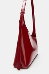 Bolso de hombro rojo de diseño asimétrico en acabado brillante. Cuenta con asa ajustable, bolsillo interior y cierre mediante cremallera metálica.