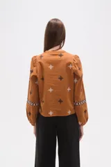 Blusa de algodón color tostado con cuello mao y escote en V. Presenta mangas largas abullonadas con puños elásticos y un diseño con bordados geométricos en contraste distribuidos por toda la prenda.