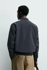 Cazadora bomber ligera de tejido técnico color verde oliva, con cuello, puños y bajo en rib elástico. Presenta cierre frontal de cremallera y bolsillos de vivo en la cadera.