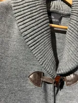 Chaleco de lana merino color gris, sin mangas, con cuello chal tejido en rib, bolsillos frontales applicados con ribete de cuero y cierre de botones tipo duffle con presillas de cuero.