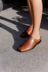 Mules de cuero color suela, con diseño abierto y tira frontal decorada con tachas metálicas circulares. Presentan punta cuadrada y suela plana.