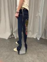 Pantalón de jean azul raw con diseño de corte versátil, ajustable mediante cierres laterales que revelan un panel interior rojo. Presenta una estampa gráfica de gran tamaño en la parte trasera con detalles bordados, costuras reforzadas y etiqueta de cuero en la cintura.