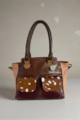 Cartera de cuero combinada en tonos marrones y beige, con detalles en animal print estilo pelo de vaca en los bolsillos frontales. Cuenta con doble asa de mano y herrajes metálicos.