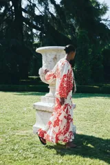 Vestido largo de tejido ligero color blanco con estampado floral rojo. Tiene cuello redondo con lazos, mangas globo largas con puños abotonados, corte holgado y falda con volantes en el ruedo.