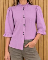 Camisa de denim y algodón color lila, con cuello alto con volados, botonadura frontal con botones oscuros, mangas 3/4 abullonadas en los hombros y terminaciones con volados en los puños.