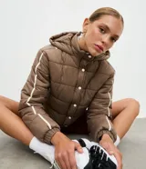 Campera puffer deportiva color marrón con capucha integrada y cierre frontal con botones a presión. Presenta diseño acolchado con bieses laterales en color blanco a lo largo de las mangas. Cuenta con puños elásticos y cuello alto.