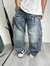Pantalón de jean con corte baggy, de lavado ácido y diseño holgado. Presenta bolsillos traseros con parches en forma de cruz y detalles bordados con motivos tribales.