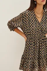 Vestido corto de escote en V cruzado, con mangas tres cuartos y cintura elástica. Presenta un diseño estampado con motivos geométricos en tonos oscuros y detalles metalizados, con un ruedo con volado.