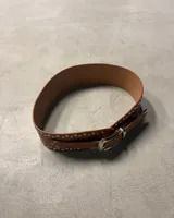 Cinturón ancho de cuero negro con doble hilera de tachas plateadas. Hebilla metálica plateada de estilo occidental y punta de cierre en forma de flecha.