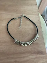 Collar corto de cuero negro con cuentas plateadas y dijes circulares colgantes.