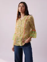 Blusa de gasa estampada con flores amarillas y verdes, marca Alex & Lili. Tiene cuello mao, mangas 3/4 acampanadas y cierre frontal con botones. Presenta volados verticales color naranja en el frente y en las mangas.