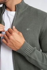 Campera tejida de punto en color verde militar, con cierre frontal completo, cuello elevado y ajuste regular. Presenta un diseño texturizado con logo bordado en el pecho y puños acanalados.