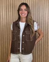 Chaleco de denim marrón con costuras a contratono en color blanco, cuello camisero, bolsillos delanteros con solapa y cierre frontal con botones metálicos.