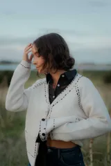 Saco tipo cardigan de punto color blanco con detalles de pespuntes decorativos en color negro a lo largo de los bordes y costuras. Presenta cuello en V, cierre frontal con botones y mangas largas.