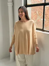 Sweater de punto color beige, de corte holgado y diseño asimétrico con tajos laterales. Presenta cuello redondo y mangas largas con puños acanalados, además de un detalle de costura vertical central.