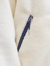 Campera de tejido tipo teddy color crema, con cuello alto, cierre frontal completo y ribetes en color azul marino. Presenta bolsillos laterales con cierre y bordado en el pecho.