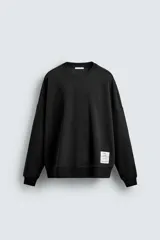 Sudadera negra de corte relaxed fit, confeccionada en tejido técnico interlock de algodón con elasticidad. Presenta cuello redondo, manga larga y una etiqueta aplicada en el bajo. Los acabados son en rib.