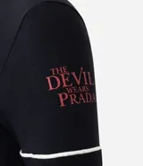 Remera negra de algodón con cuello redondo y mangas cortas con ribete blanco. Presenta un estampado frontal con motivos gráficos inspirados en la película El Diablo Viste a la Moda, incluyendo recortes tipo post-it, ilustraciones y texto.