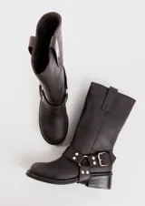 Bota de caña media confeccionada en cuero vacuno color marrón oscuro, con diseño de estilo biker. Presenta una tira decorativa con hebilla metálica y estribo desmontable en la zona del tobillo, taco cuadrado de madera y suela de caucho.