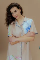 Vestido corto beige de lino y algodón, con cuello camisero con solapa, mangas cortas, cierre frontal con botones y detalles de bordado artesanal en los bordes. Presenta estampado botánico con flores y insectos.