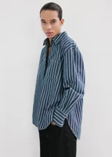 Camisa oversize de algodón estilo denim, de color azul con estampado de rayas, cuello camisero, manga larga con puños abotonados, bolsillo de parche en el pecho y cierre frontal con botones.